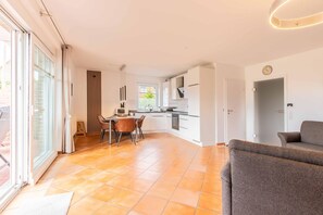 Eine moderne Küche mit weißen Schränken, ein Essbereich mit Tisch und Stühlen und ein graues Sofa in einem hellen Wohnraum mit großen Fenstern.