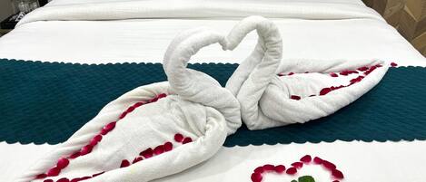 A neatly made bed with a towel arranged in the shape of swans, a heart made of rose petals, and teal pillows.