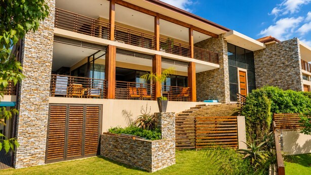 A modern two-story house with a stone facade, a balcony with wooden railings, and a well-maintained lawn. - The Cozy Chartwell Spec (uMhlanga)