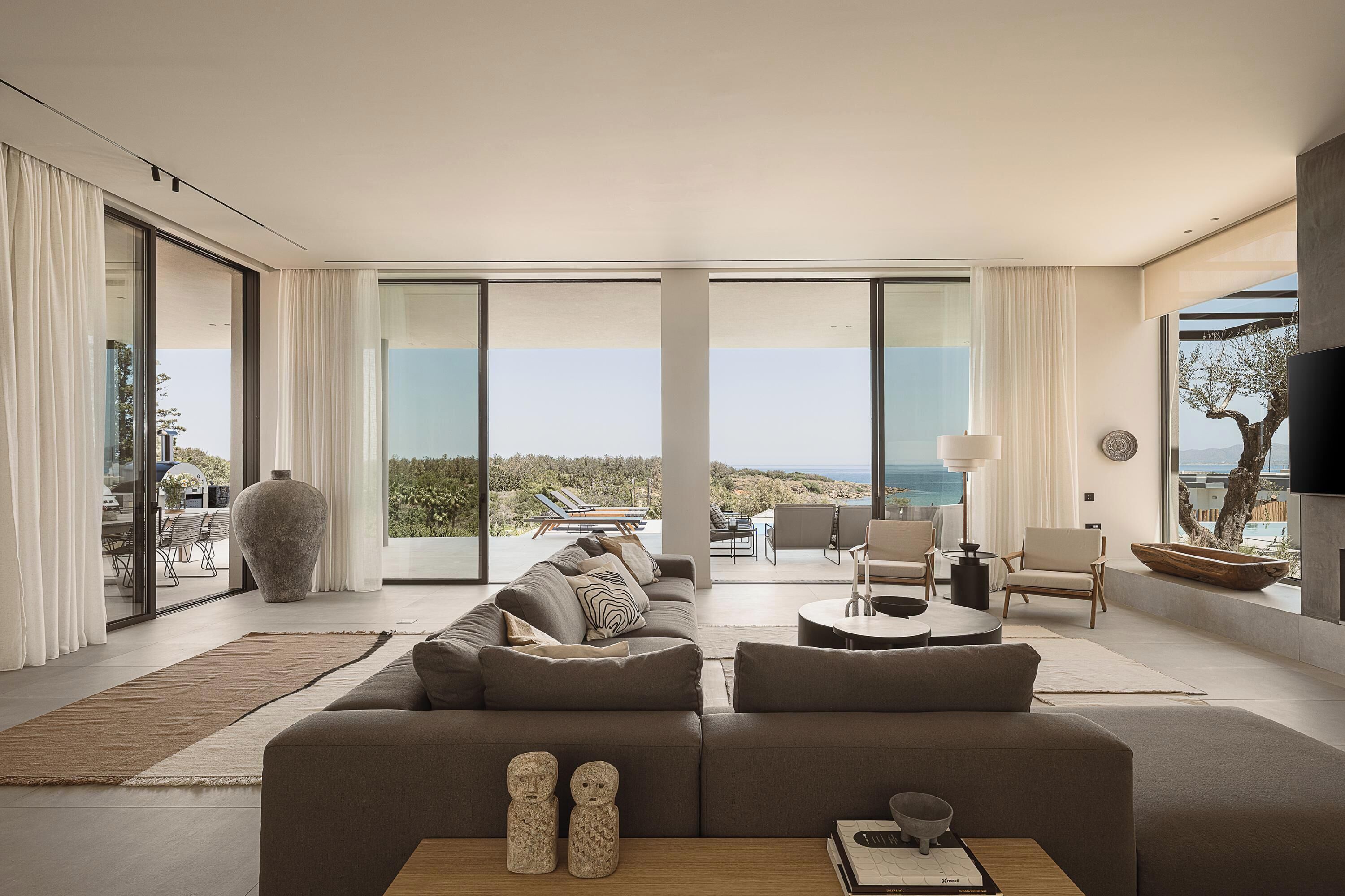 A modern living room with a large sofa, a coffee table, and a TV. There is a view of the outdoors through large windows.
