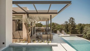 Un área de piscina moderna al aire libre con una zona de estar cubierta, sillas de descanso y vistas del paisaje circundante.