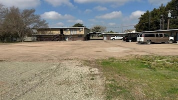Un estacionamiento amplio con un edificio al fondo, una zona techada y varios vehículos.