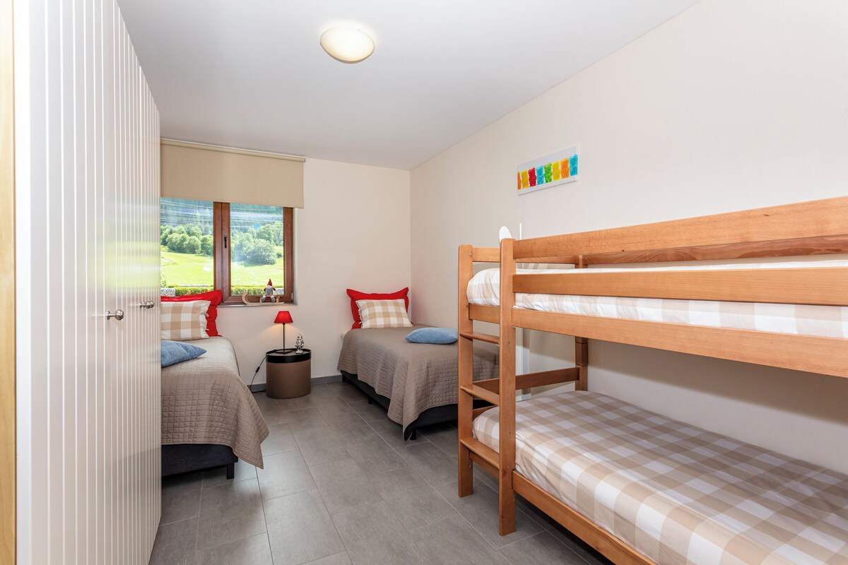 Quarto com beliches, janela com vista para vegetação, mesa de cabeceira com abajur e quadro fixado na parede.