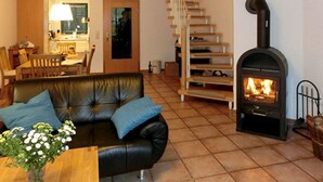 A living room with a black leather sofa, a wooden coffee table with a vase of flowers, a fireplace, a staircase, and a kitchen area in the background. - Malve 3 Thatched-Roof House - Malve 3 Vacation Home – Thatched-Roof Vacation with a Fireplace and Wallbox (Poseritz)