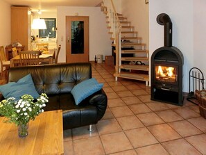 A living room with a black leather sofa, a wooden coffee table with a vase of flowers, a fireplace, a staircase, and a kitchen area in the background. - Malve 3 Thatched-Roof House - Malve 3 Vacation Home – Thatched-Roof Vacation with a Fireplace and Wallbox (Poseritz)