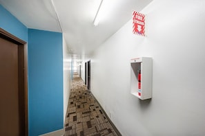 A hallway with a fire extinguisher, a fire exit sign, and a patterned carpet. - King Bed | Non-Smoking (Portland OR)