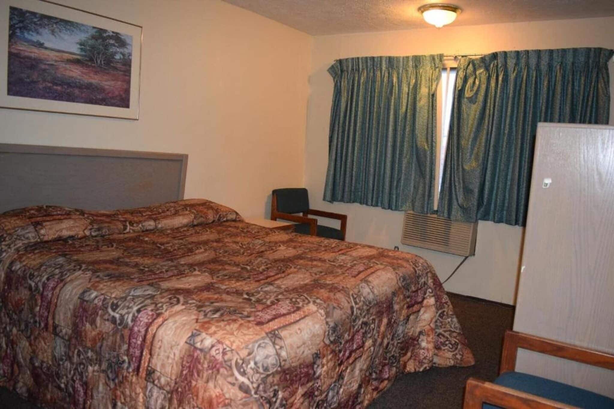 A hotel room with a bed, a chair, a desk, a window with curtains, and a wall-mounted air conditioning unit.