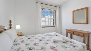 A bedroom with a bed, a nightstand with a lamp, a wooden dresser, a window with curtains, and a mirror on the wall. - Charming Bungalow Rental in Abades, Spain - Perfect Family Retreat (Abades)
