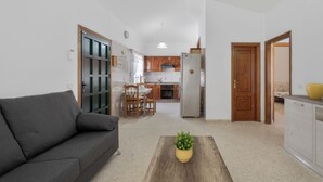 A living room with a sofa, a coffee table with a plant, a kitchen in the background, and a wooden door. - Charming Bungalow Rental in Abades, Spain - Perfect Family Retreat (Abades)