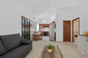 A living room with a sofa, a coffee table with a plant, a kitchen in the background, and a wooden door. - Charming Bungalow Rental in Abades, Spain - Perfect Family Retreat (Abades)