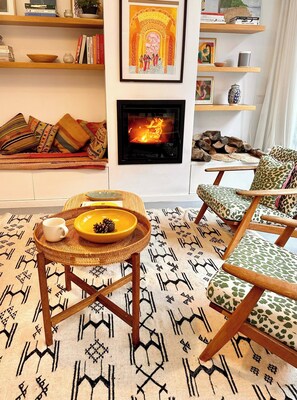 Ein gemütliches Wohnzimmer mit Kamin, einem gemusterten Teppich, einem Couchtisch mit Schale und Tasse sowie einem Sessel mit dekorativer Polsterung.