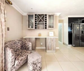 A living room with a sofa, ottoman, and a kitchen area with cabinets and a refrigerator.