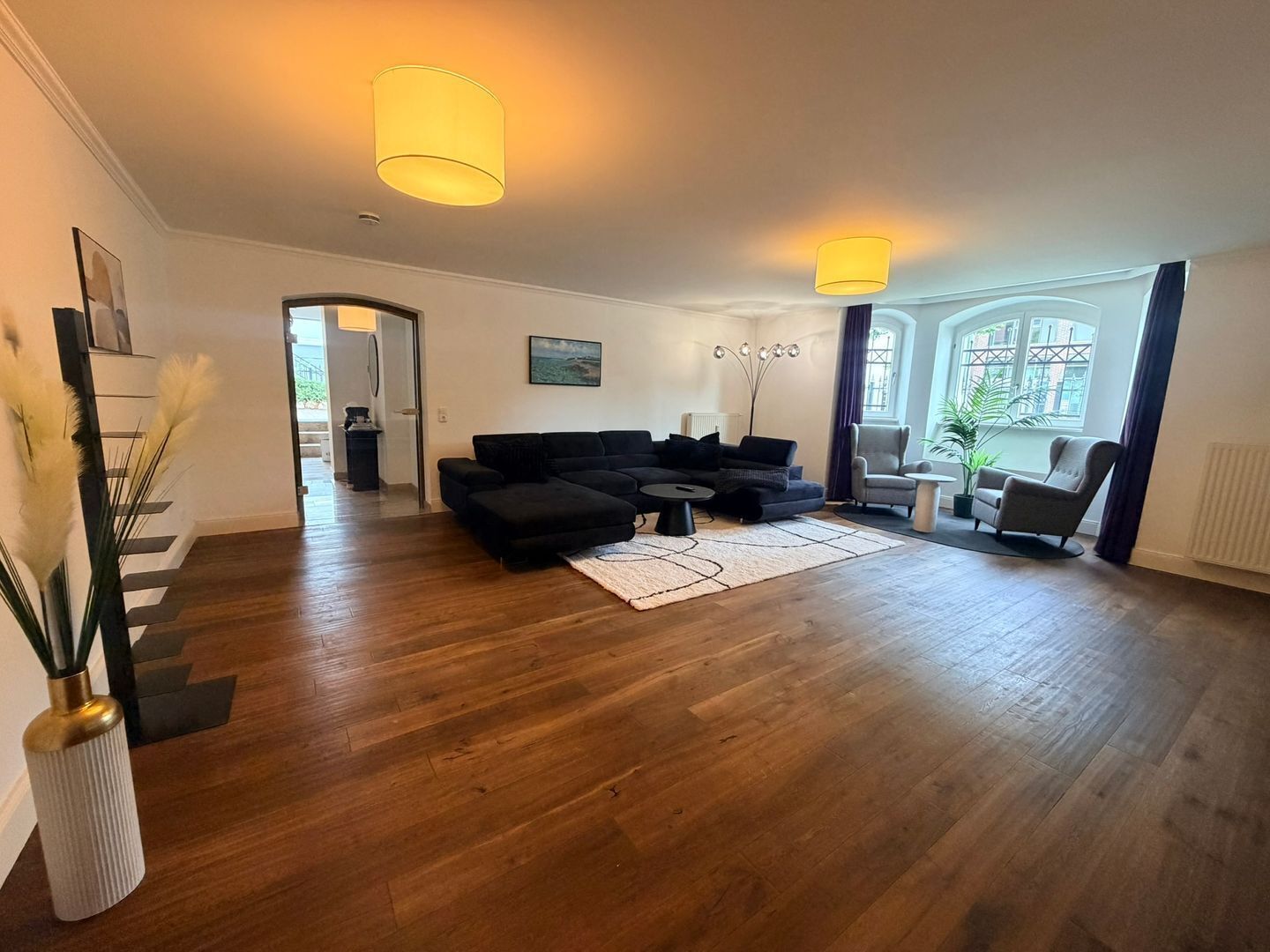 Ein geräumiges Wohnzimmer mit Holzdielenboden, einem schwarzen Modulsessel, einem Couchtisch und zwei Pendelleuchten.