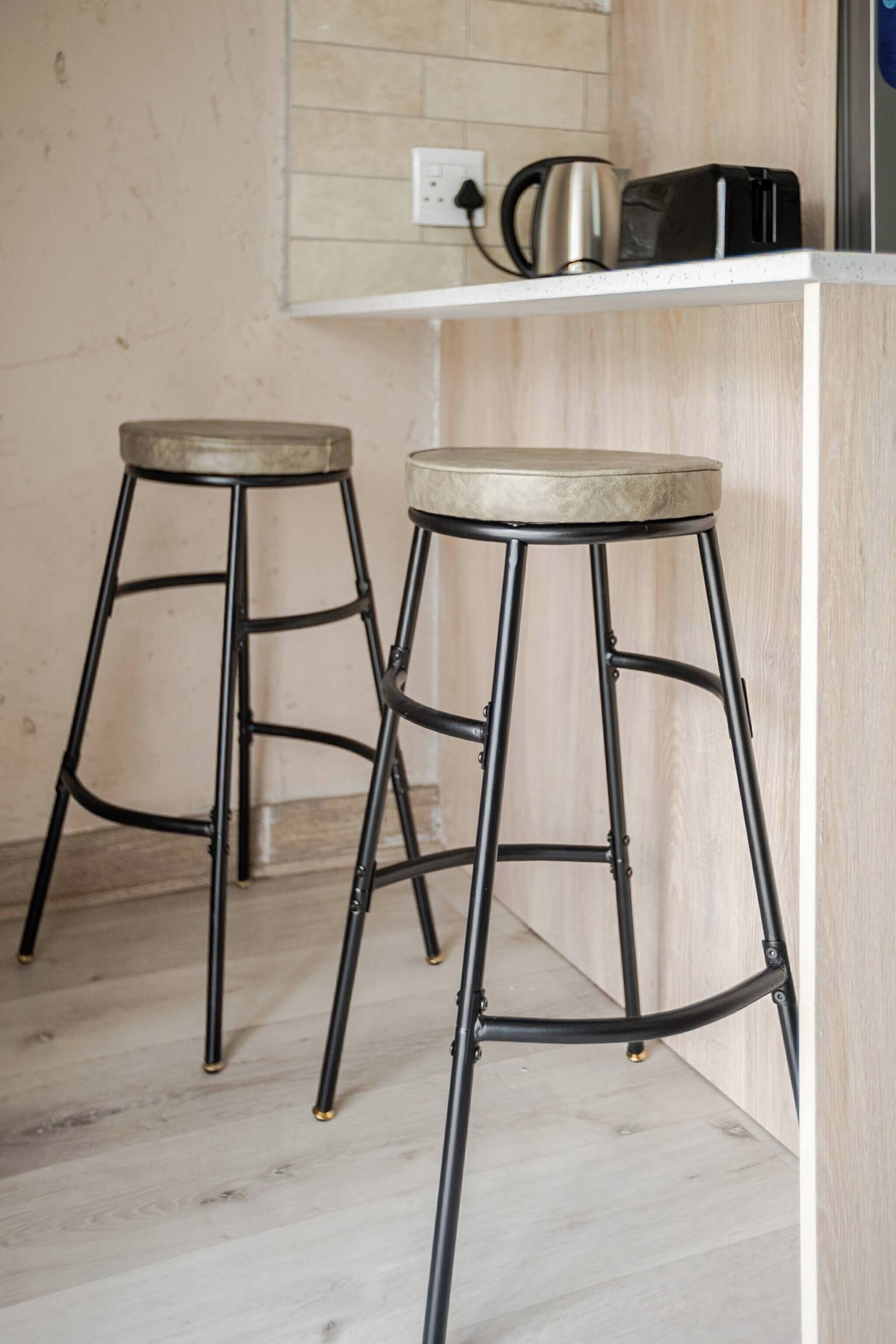 Zwei Metallhocker mit gepolsterten Sitzen, eine weiße Ablagefläche mit Wasserkocher und Toaster sowie ein heller Holzboden.