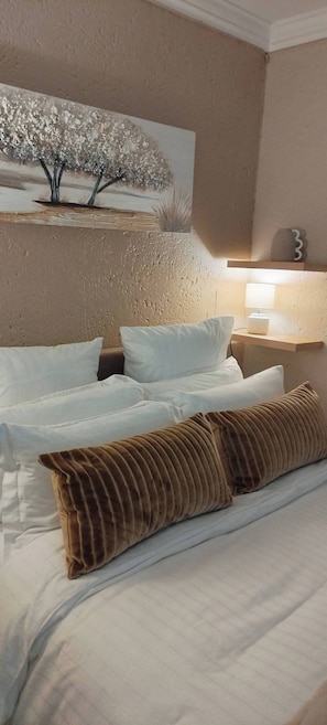 Ein Bett mit weißer Bettwäsche und einem braunen Kopfkissen, ein Wandbild, eine Nachttischlampe und ein kleines Regal mit einer Dekoration.