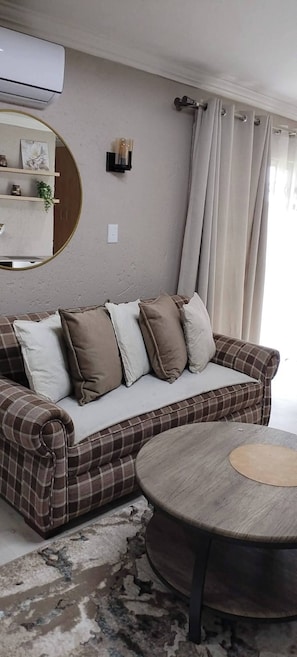 Ein Wohnzimmer mit einem karoüberzogenen Sofa, einem runden Holztisch, einer Wand-Klimaanlage und einem Spiegel.