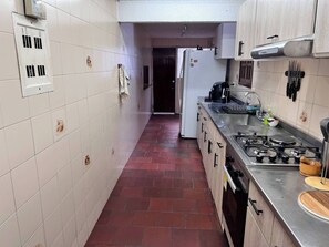 Eine schmale Küche mit weißen Fliesenwänden, einem roten Backsteinboden, einem Edelstahlherd und einem Kühlschrank.