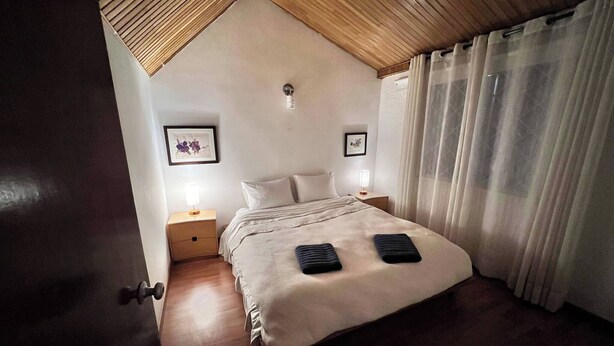 A bedroom with a wooden ceiling, a bed with two pillows, two bedside tables with lamps, and a window with curtains. - The House at north of Bogota (Bogotá)