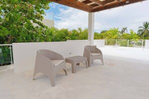 Eine Terrassenanlage im Freien mit zwei Korbstühlen und einem kleinen Tisch, umgeben von üppiger Bepflanzung und einer weißen Mauer.