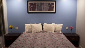 A bedroom with a patterned bedspread, two bedside tables, a framed artwork on the wall, and a vase with flowers on each side of the bed.