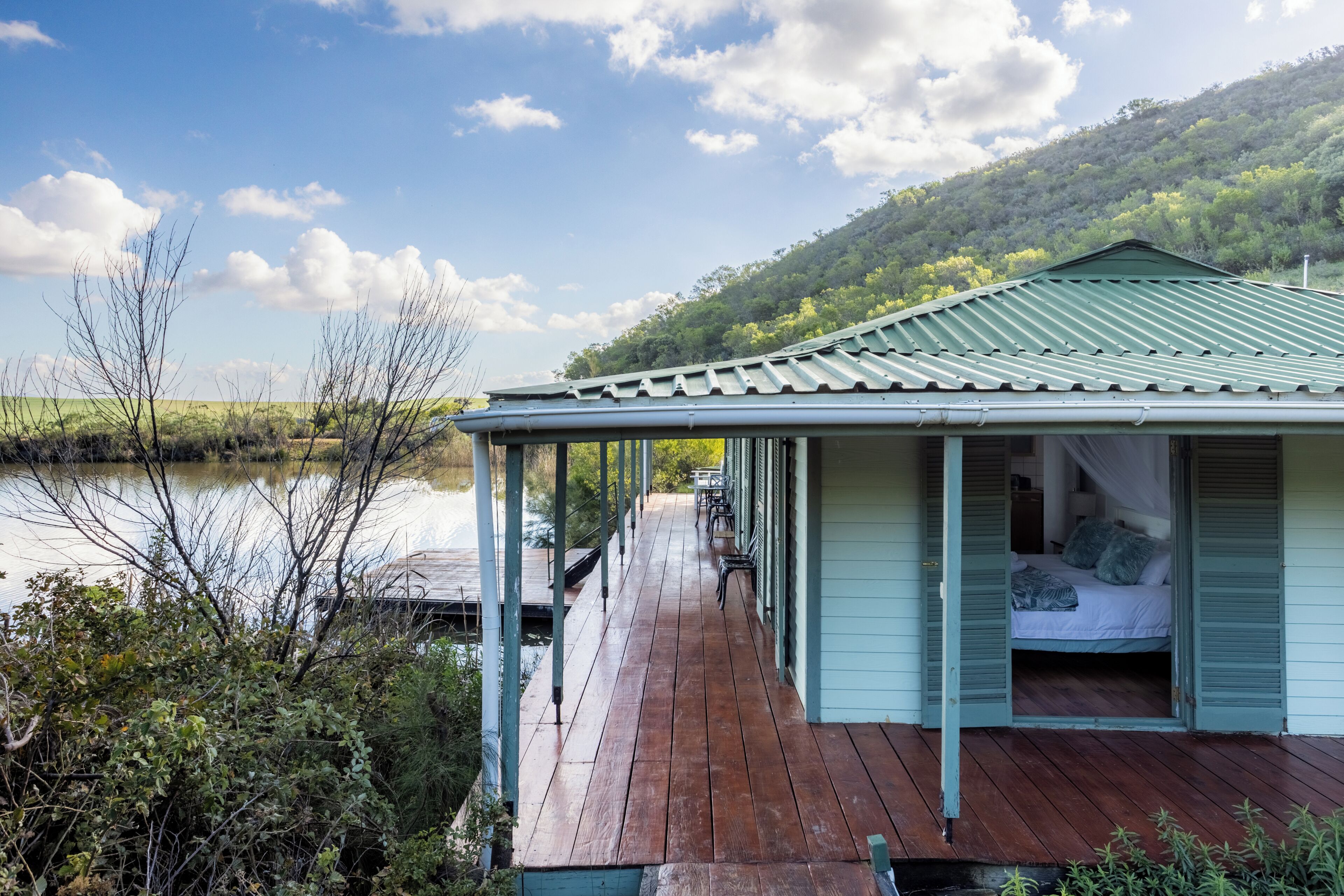 A wooden deck leading to a building with a green roof, overlooking a calm body of water and a hilly landscape.