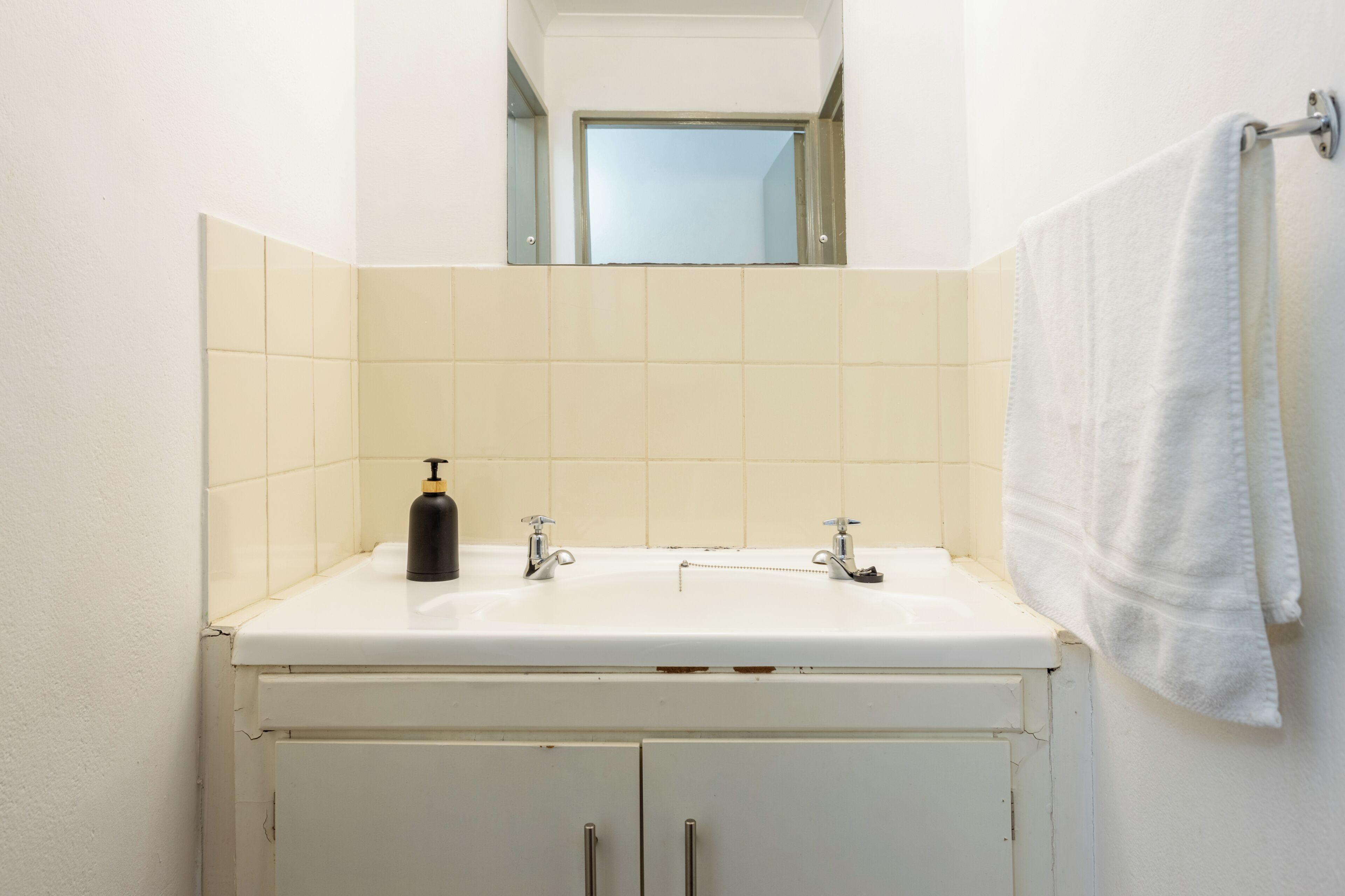 Un baño con un lavamanos blanco, dos grifos, un espejo sobre el lavamanos y una toalla colgada en un perchero.