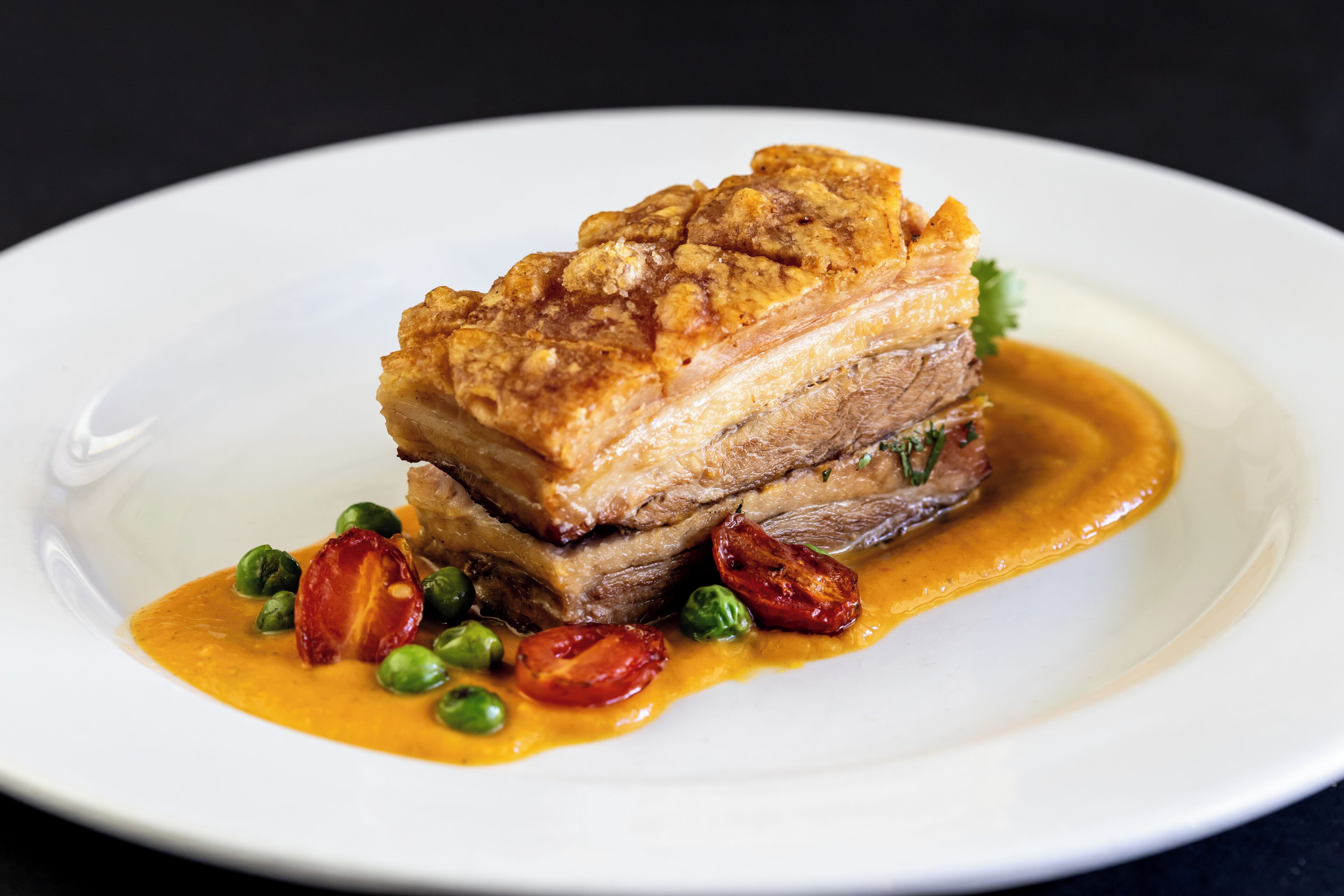 A plate with a pork dish, possibly a pork belly, accompanied by peas and cherry tomatoes, and a sauce.
