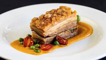 A plate with a pork dish, possibly a pork belly, accompanied by peas and cherry tomatoes, and a sauce.