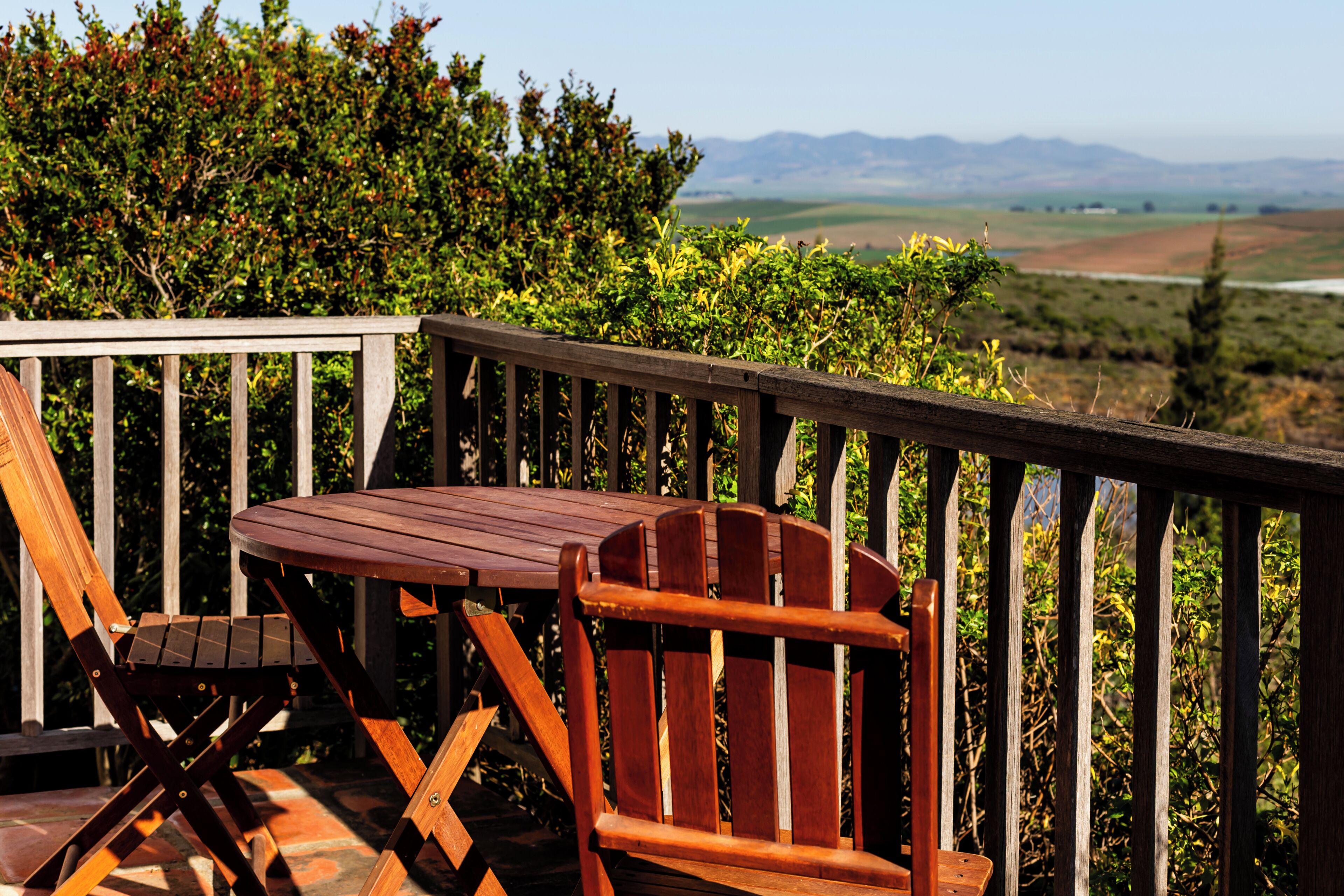 Terraza de madera con una mesa y dos sillas, con vistas a un paisaje de montañas y vegetación.
