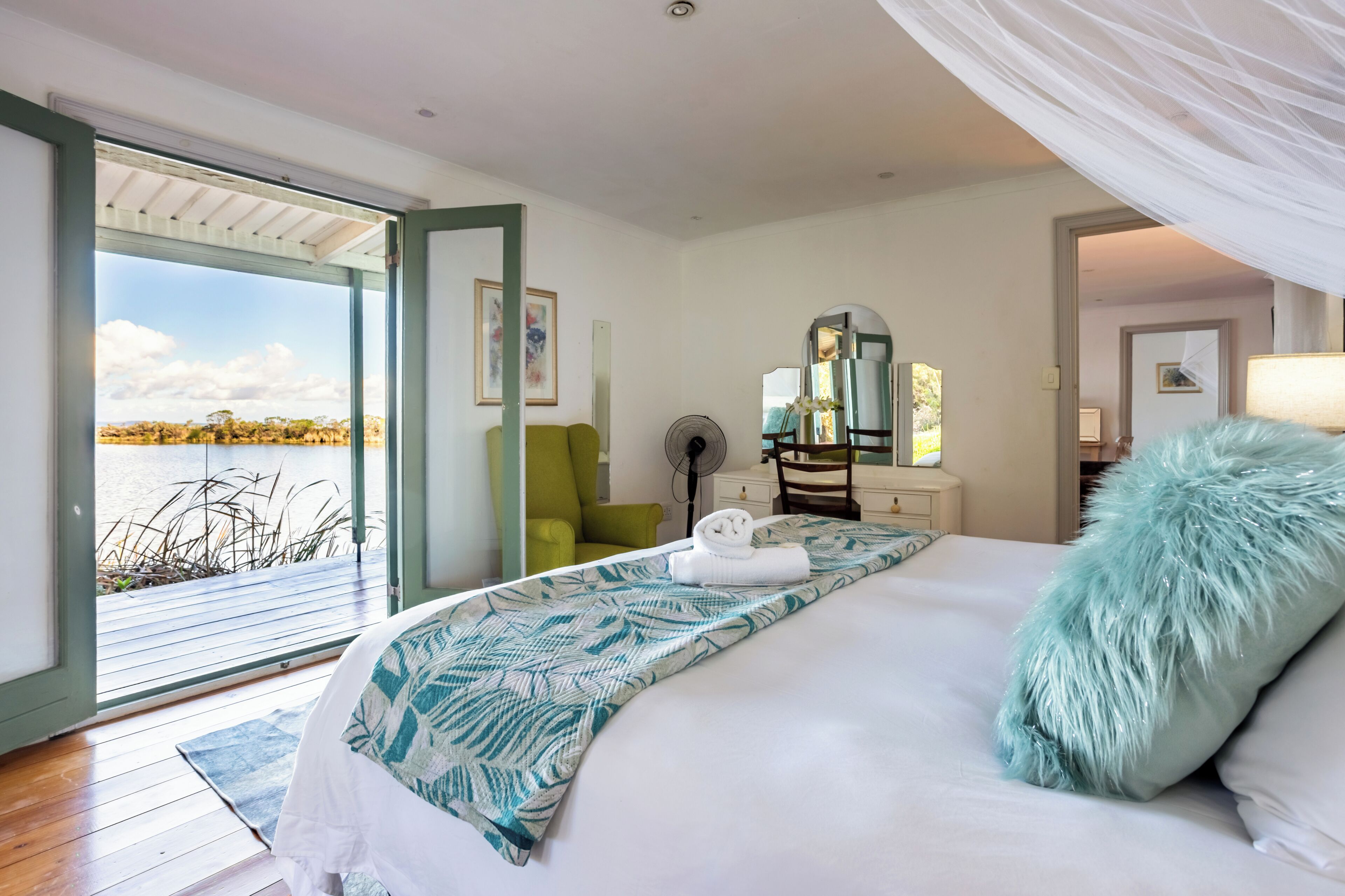 A bedroom with a bed, a green chair, a fan, and a view of a body of water.