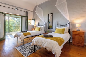 A bedroom with two beds, a wooden floor, a patterned rug, and a view of greenery outside.
