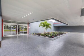 Entrada de um edifício moderno com porta de vidro, piso de concreto e um pequeno jardim com palmeiras.