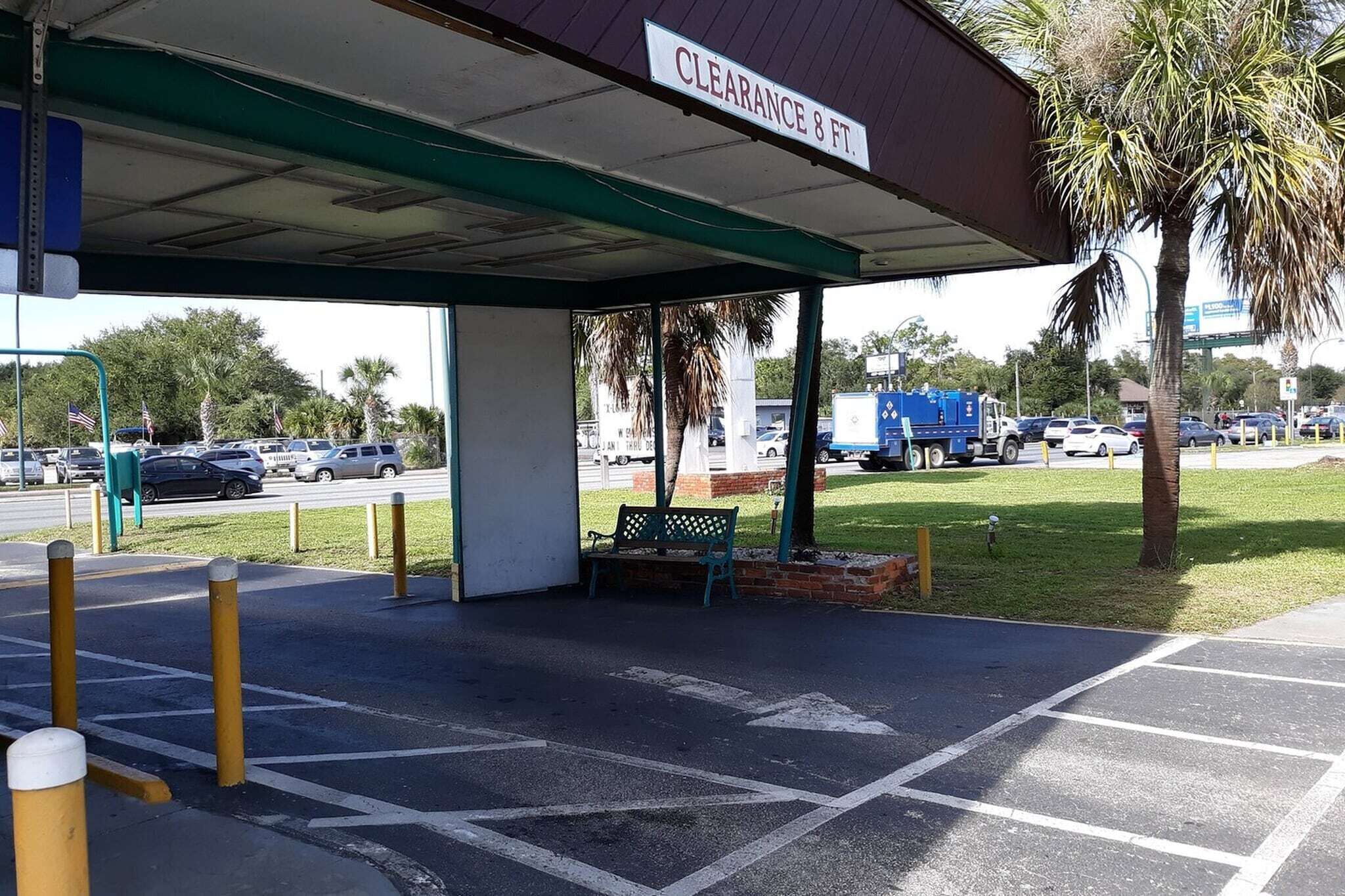 A gas station with a sign reading 'CLEARANCE 8 FT.' There is a bench under the canopy, and the area is surrounded by palm trees and a parking lot with cars.