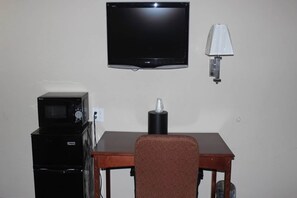 A room with a television mounted on the wall, a microwave, a lamp, and a table with a chair.