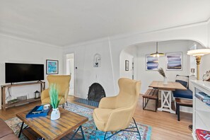 Ein Wohnzimmer mit Fernseher, zwei Sesseln, einem Couchtisch, einem Kamin und einer Essecke mit Tisch und Bank.
