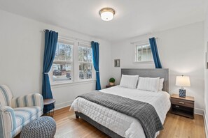 Ein Schlafzimmer mit einem Bett, einem Nachttisch, einer Lampe, einem Sessel, einem kleinen Tisch und einem Fenster mit Vorhängen.