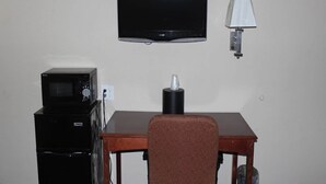 Una habitación con televisión en la pared, microondas, lámpara y una mesa con una silla.