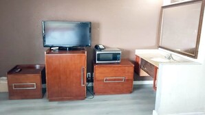 Una habitación compacta con un armario de madera, un televisor, un microondas, un cajón pequeño y un espejo.
