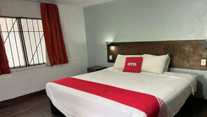 Une chambre d’hôtel comprenant un lit, une table de chevet, une fenêtre avec des rideaux et une tête de lit dotée d’un luminaire.
