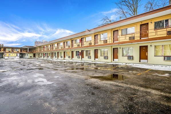A row of motel rooms with individual doorways and windows, a parking lot with puddles, and a clear sky.