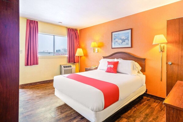 A hotel room with a large bed, two bedside lamps, a window with curtains, an air conditioning unit, and a wooden door.