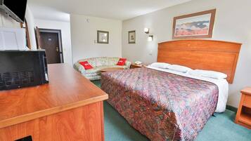 Quarto de hotel com escrivaninha de madeira, cama com colcha estampada, sofá floral com almofadas vermelhas e um quadro emoldurado na parede.
