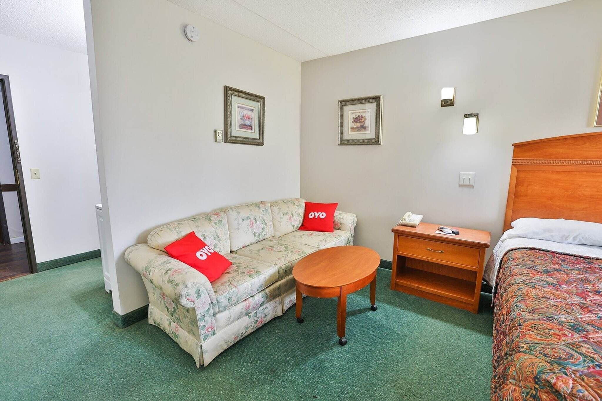 A hotel room with a floral sofa, a round wooden table, a bed with a patterned bedspread, a nightstand, and two framed pictures on the wall.