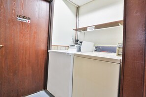 Une pièce privée avec une porte en bois, un lave-linge blanc et une étagère sur laquelle se trouvent une machine à café et une boîte étiquetée « Lave-linge industriel Good Earth ».