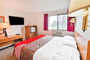 A hotel room with a bed, a desk with a lamp, a TV, a wardrobe, and a window with curtains. - Pipestone|Queen Bed|Nsmk (Pipestone)