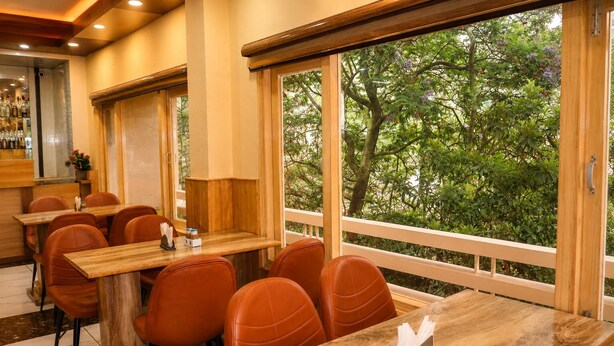 A restaurant with wooden tables and chairs, a bar area, and large windows with a view of trees. - Jannat Hotel - Solan (Solan)