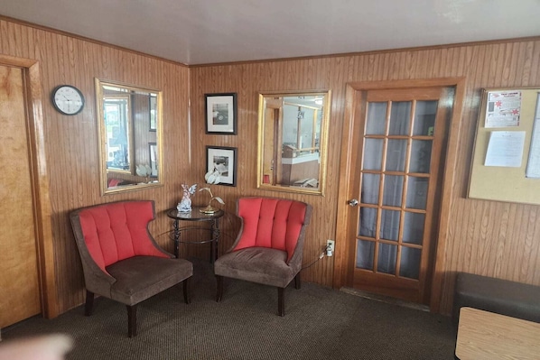 A room with wooden paneled walls, two red upholstered chairs, a small round table with a lamp, and a door with a frosted glass panel.