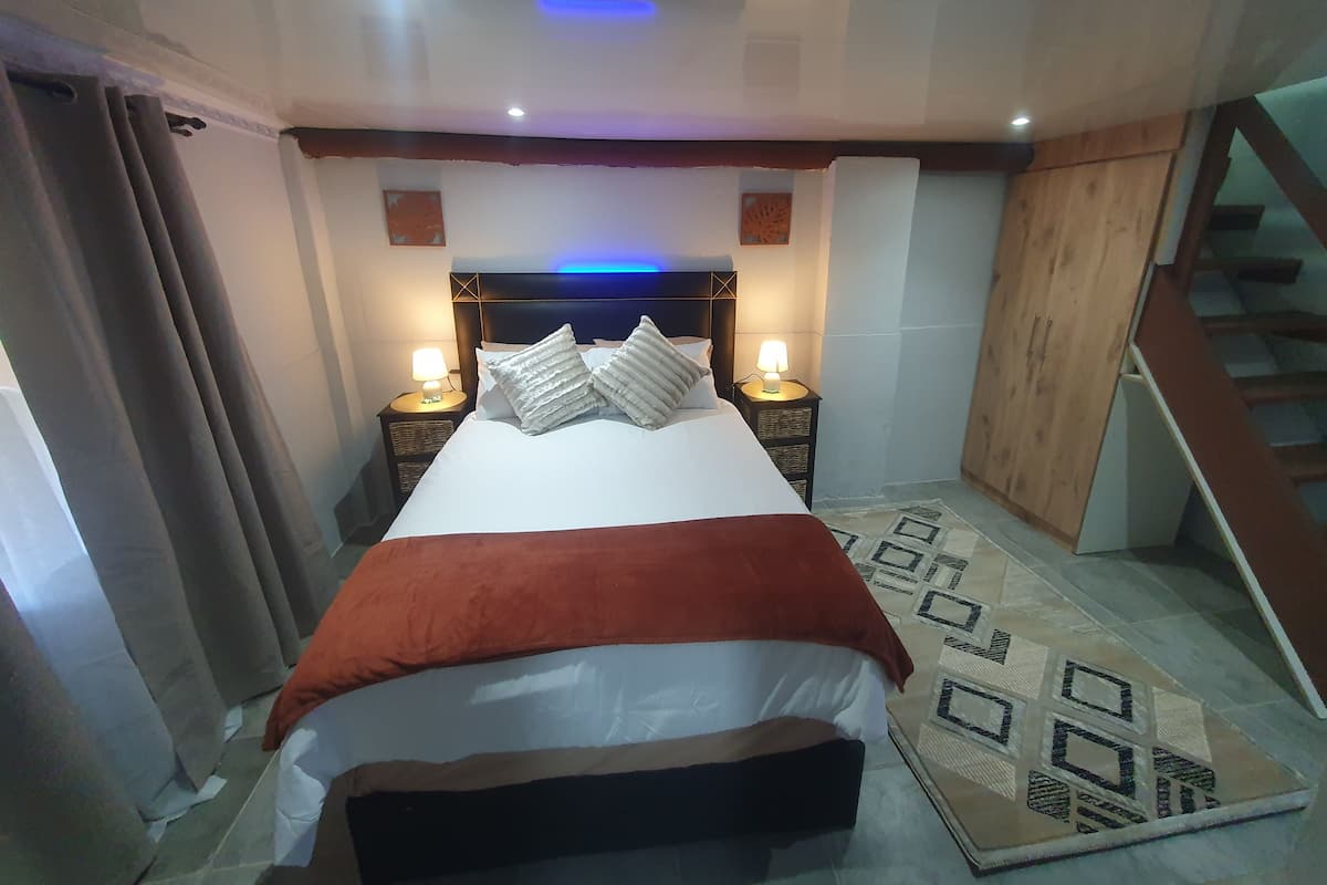 Une chambre à coucher avec un lit, deux tables de chevet avec des lampes, une tête de lit, un escalier en bois et un tapis à motifs.