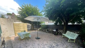 A gravel patio with a table and chairs, a large umbrella, a grill, and a bench. - Le Puy d’Eraigne: peaceful, cool apartment with terrace and optional garage on request (Saint-Nectaire)