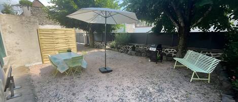 A gravel patio with a table and chairs, a large umbrella, a grill, and a bench.